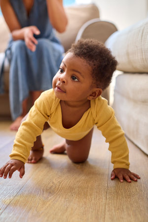 Smiling Baby Girl Learning To Crawl On Floor At Home With Mother Encouraging In Backgroundの写真素材
