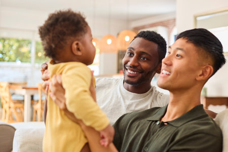 Same Sex Male Couple Or Friends Playing With Baby Sitting On Sofa At Home Together