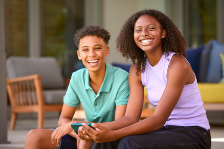 Portrait Of Teenage Brother And Sister With Mobile Phone Sitting Outdoors At Home On Deck