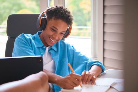 Teenage Boy Sitting At Desk In Bedroom At Home Doing Homework Streaming Music To Headphonesの写真素材