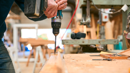 Close Up Of Carpenter At Workbench In Workshop Using Cordless Electric Drill