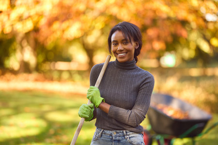 Portrait Of Young Woman With Barrow Raking Leaves In Autumn Garden