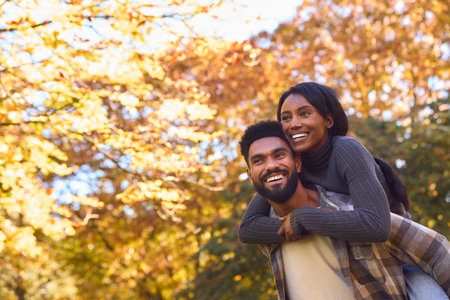 Loving Young Couple With Man Giving Woman Piggyback Against Background Of Autumn Leavesの写真素材