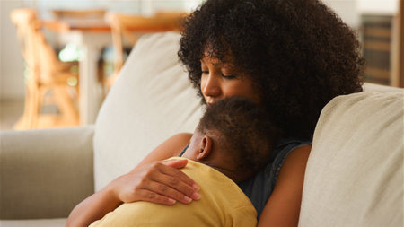 A mother embraces her baby tenderly while sitting on a comfortable sofa in a warmly lit living room. The child rests peacefully against her, creating a moment of bonding and affection.の写真素材