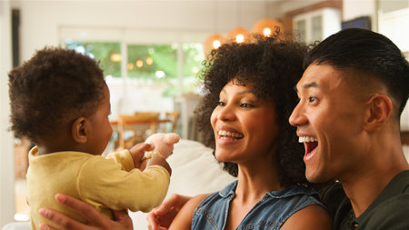 A young couple is joyfully interacting with their adorable baby in a warm and inviting living room. The parents display wide smiles while the baby reaches out playfully, creating a heartwarming atmosphere filled with love and connection.の写真素材