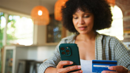 A young woman sits in a cozy indoor setting, intently using her smartphone while holding a credit card. The room is warmly lit with hanging pendant lights, and she appears focused on completing an online purchase.の写真素材