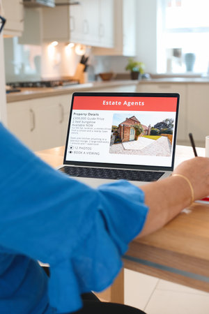 A person is focused on their laptop, exploring property details for real estate in a modern kitchen....