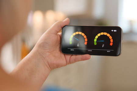 A person is examining a smartphone that showcases an energy consumption meter with colorful indicators. The setting is bright and modern, suggesting a focus on energy efficiency. The interface displays various readings crucial for monitoring electricity usage and water consumption.の写真素材
