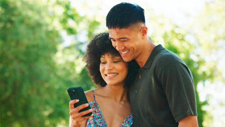 A couple smiles as they look at a smartphone in a bright outdoor setting.