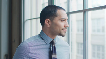 Head And Shoulders Portrait Of Smiling Young Businessman Looking Out Of Window In Office
