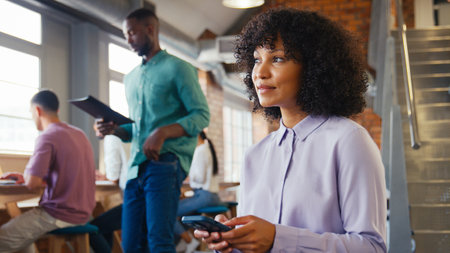 Smiling Businesswoman Using Mobile Phone Working In Open Plan Office With Colleagues In Background