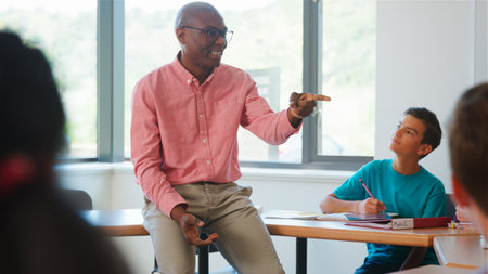 Male High School Tutor Sitting On Desk Teaching Group Of Multi-Cultural Students In Classroom