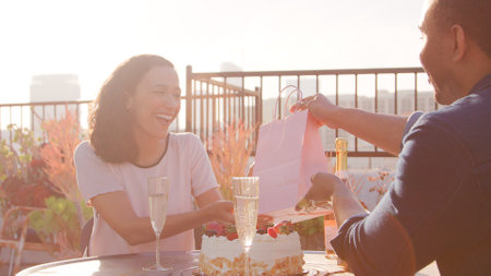 Man Giving Woman Gift As They Celebrate On Rooftop Terrace With City Skyline In Backgroundの写真素材