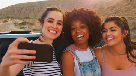 Three Female Friends Sitting In Trunk Of Open Top Car Taking Selfie On Road Trip Through Countryside