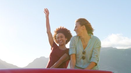 Two Female Friends Standing Up In Back Of Open Top Hire Car On Summer Vacationの写真素材