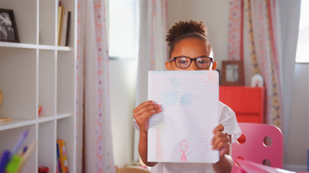Portrait Of Young Girl Wearing Glasses In Bedroom At Home Holding Up Drawing To Camera
