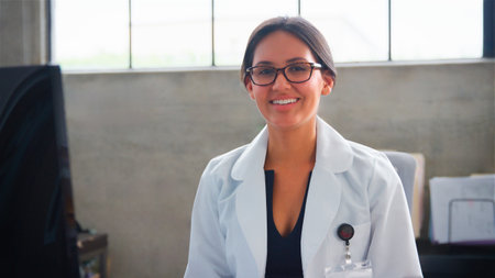 Portrait Of Smiling Female Doctor Wearing White Coat Sitting Behind Desk In Officeの写真素材