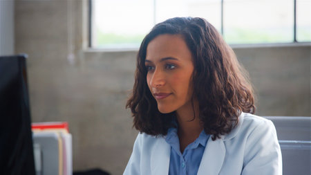 Female Doctor Wearing White Coat Sitting Behind Desk In Office Working At Computer