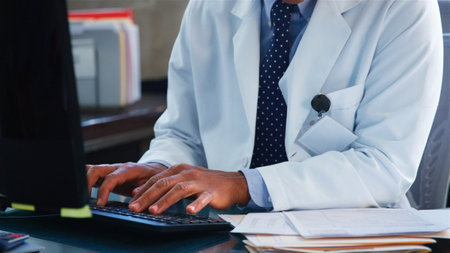 Close Up Of Male Doctor Wearing White Coat Behind Desk In Office Working At Computer