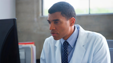 Male Doctor Wearing White Coat Sitting Behind Desk In Office Working On Computerの写真素材