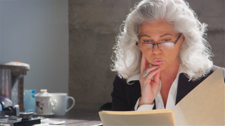 Mature Businesswoman Wearing Glasses Sitting At Desk In Office Reading Document Or Fileの写真素材