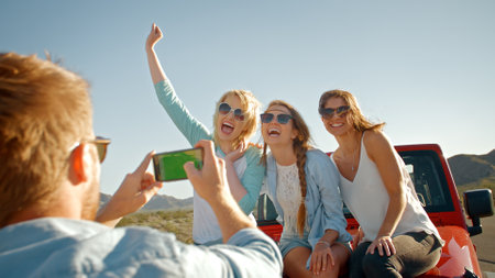 Young Man With Mobile Phone Taking Photo Of Three Women On Road Trip Sitting On Hood Of Car