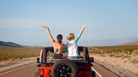 Rear View Of Friends On Road Trip Driving Convertible Car On Country Road With Women Sitting In Back