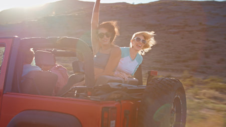 Friends On Road Trip Driving Convertible Car On Country Road With Women Sitting In Back Waving