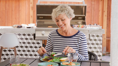 Senior Woman Outdoors At Home Sitting At Table Eating Barbecue Meal With Saladの写真素材