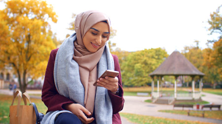 British Muslim Woman Sitting On Bench Using Mobile Phone To Check Messages And Social Media In Parkの写真素材