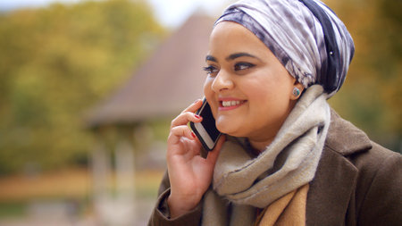 Close Up Of British Muslim Woman Sitting On Bench Talking On Mobile Phone In Autumn Park