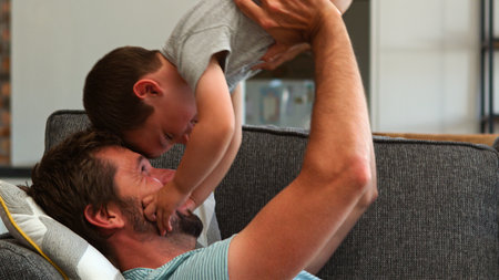 Father Lying On Sofa At Home Having Fun With Son Lifting Him Into The Airの写真素材