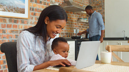 Mother And Baby Daughter Use Laptop As Father Prepares Meal At Homeの写真素材