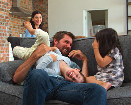 Mother Working On Laptop Whilst Father And Children Play On Sofa Togetherの写真素材