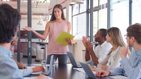 Young Businesswoman Making Presentation To Colleagues Using Laptops Sitting Around Boardroom Table