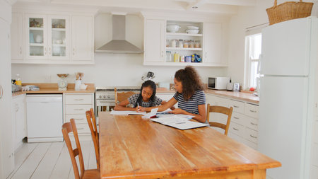 Teenage Girl Helping Sister With Homework Sitting At Table In Kitchen At Homeの写真素材
