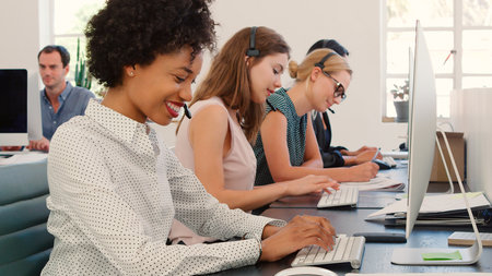 Multi-Cultural Female Business Team Wearing Headsets Working In Busy Customer Service Departmentの写真素材