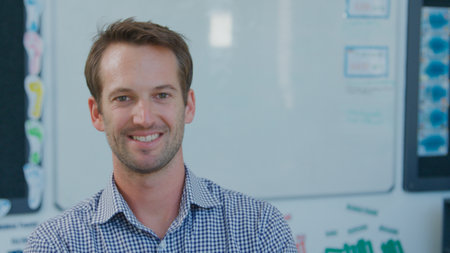 Portrait Of Smiling Male Teacher Standing In Front Of Whiteboard In Classroom
