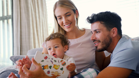 Family Sitting On Sofa At Home With Parents Reading Book With Young Sonの写真素材