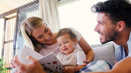 Family Sitting On Sofa At Home With Parents Reading Book With Young Son