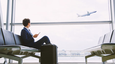 Young Businessman Sitting In Airport Departure Lounge Using Mobile Phone Watching Plane Take Offの写真素材