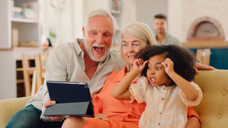 Granddaughter And Grandparents On Sofa At Home Streaming Horror Show On Digital Tabletの写真素材