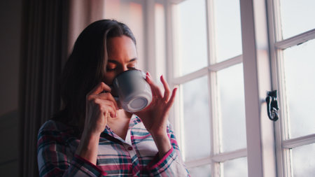 Woman At Home Looking Out Of Window Drinking Hot Drink From Mug