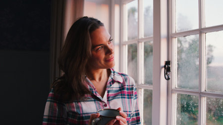 Woman At Home Looking Out Of Window Holding Hot Drinkの写真素材