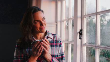 Woman At Home Looking Out Of Window Holding Hot Drinkの写真素材