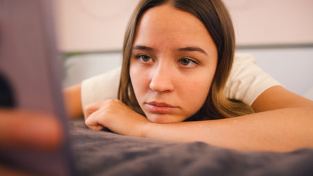 Depressed Teenage Girl Lying On Bed At Home Looking At Mobile Phoneの写真素材