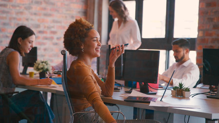 Businesswoman In Office Working On Laptop At Desk Talking Into Mic Of Mobile Phone