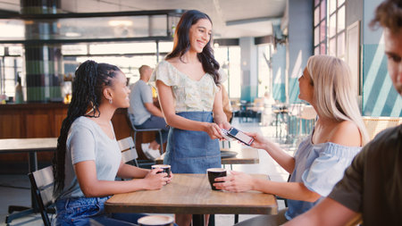 Woman Meeting Friend In Coffee Shop Paying Bill With Contactless Mobile Phone Payment