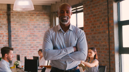 Portrait Of Smiling Mature Businessman Standing In Busy Office With Colleagues Working In Background