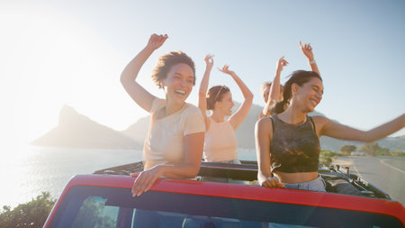Female Friends Standing Up Through Sun Roof Of Car Dancing On Road Trip